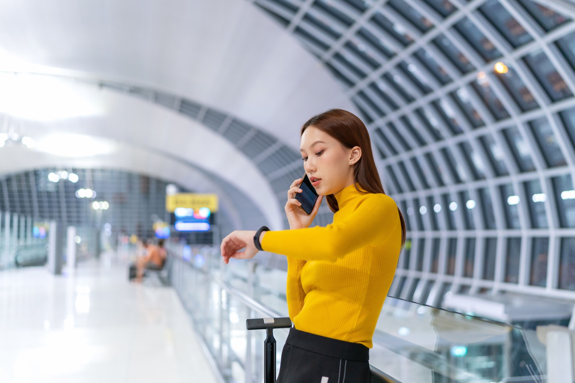 Young Asian woman talking on phone and checking time at the airport departure terminal, a suitcase by her side. Young asian businesswoman on business trip. Young Woman business on the move. Digital banking anytime, anywhere.