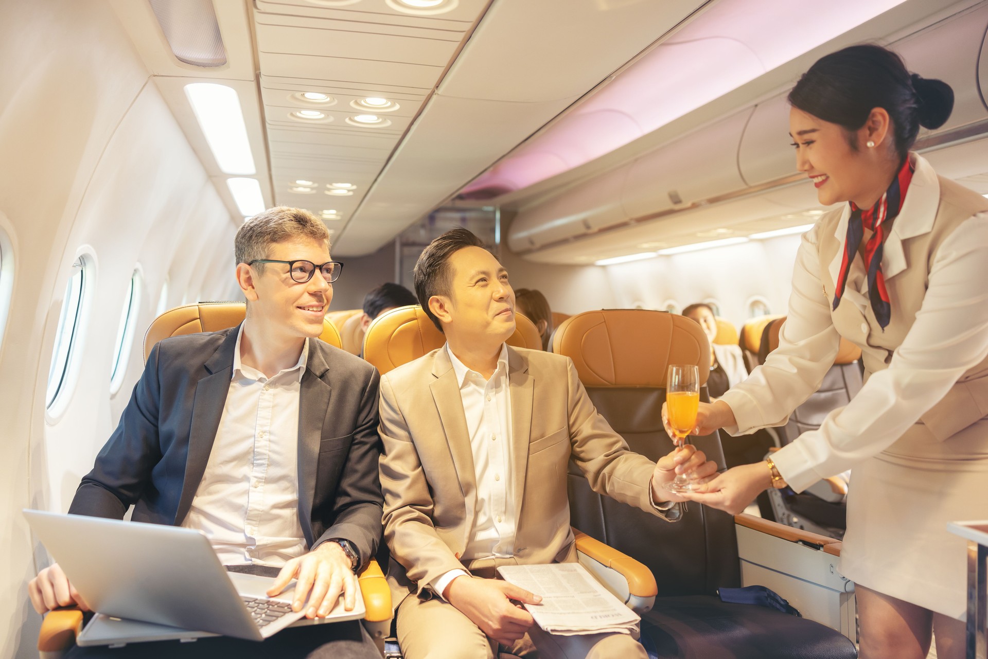 On a corporate aircraft, an air hostess serves passengers. On an airplane, a flight attendant serves a drink to a businessman who is working on his laptop.