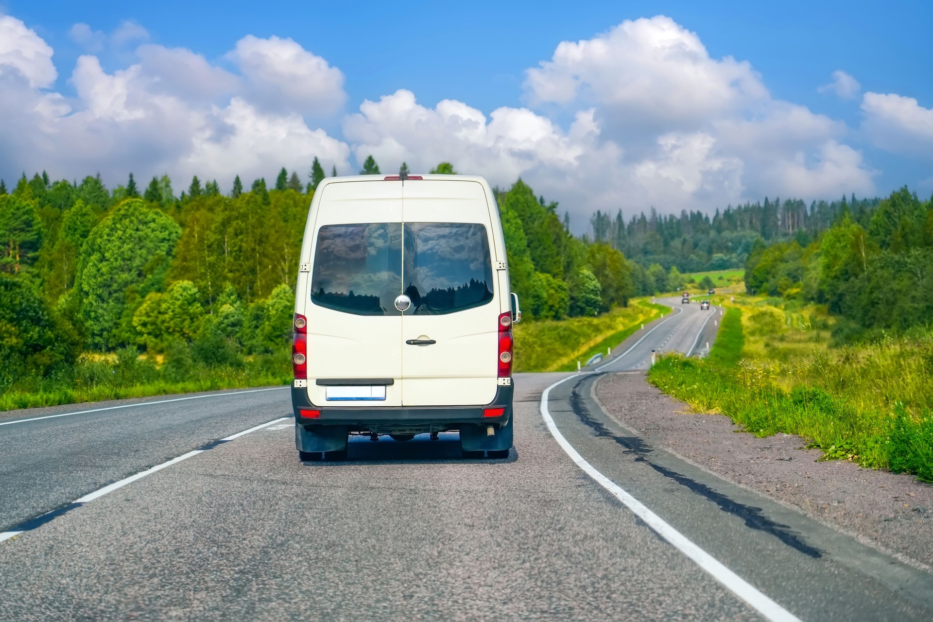 Minibus drives along a winding road in the countryside surrounded by forests on a summer day.