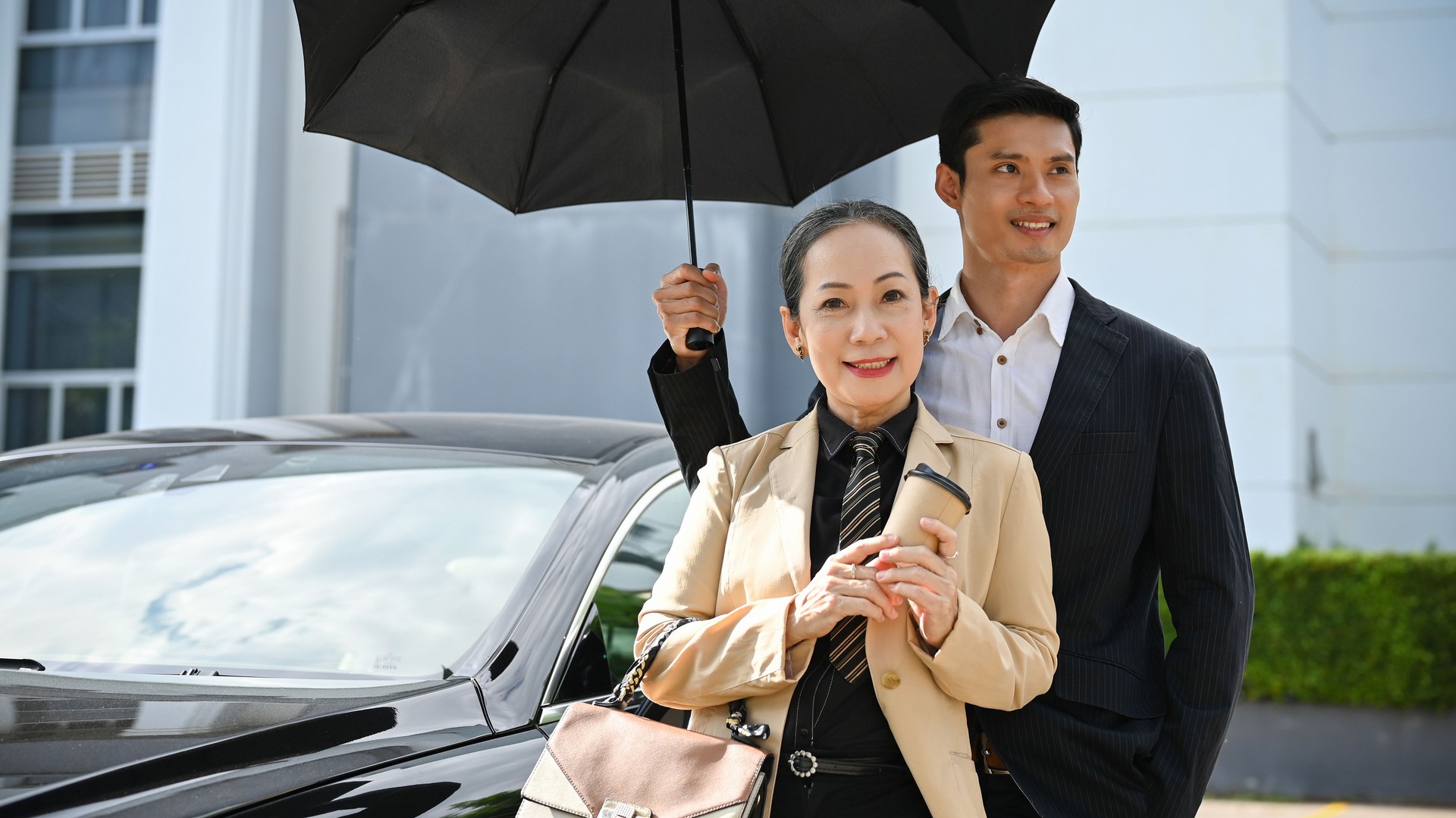 Successful Asian aged businesswoman with handsome male bodyguard under the umbrella.